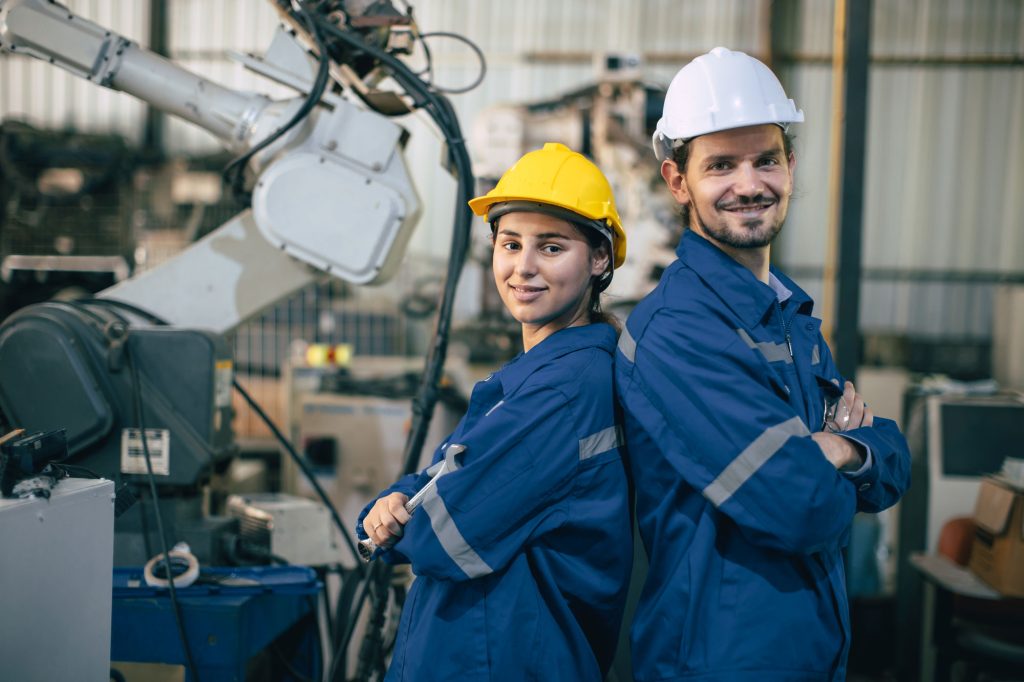 couple-worker-happy-teamwork-standing-together-friendly-smiling-enjoy-working-heavy-metal-industry-2 Two manufacturing workers standing back-to-back in front of industrial machinery, symbolizing the skilled and reliable workforce provided by Workvantage.