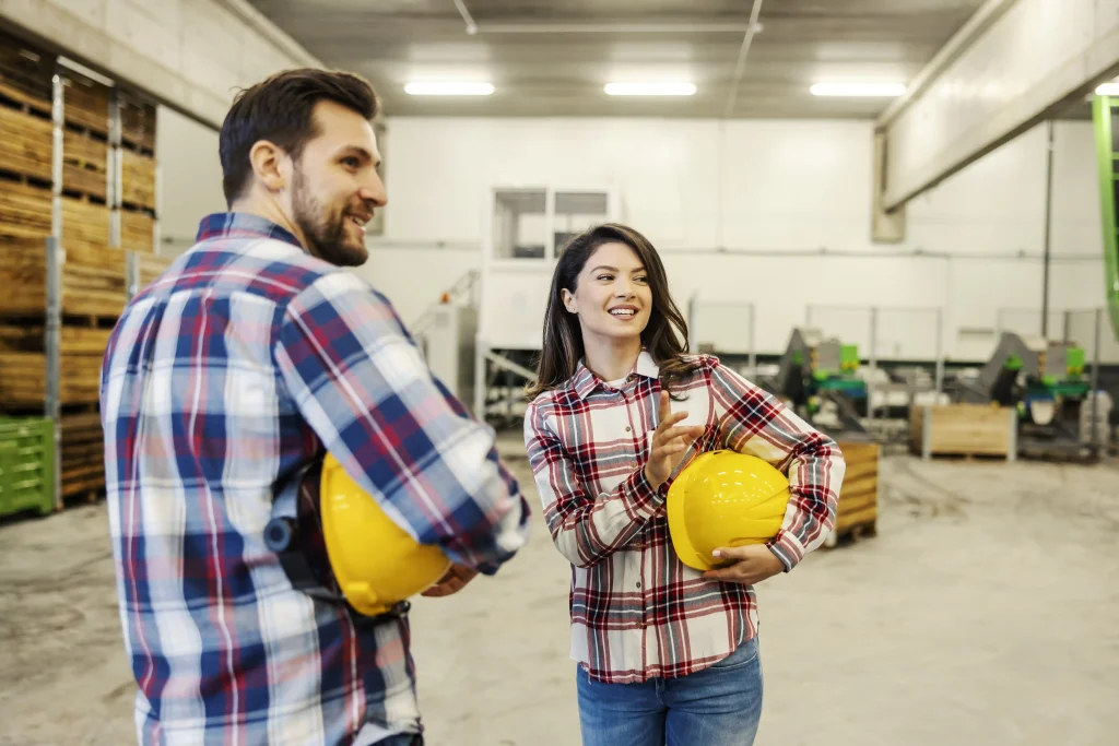 Two skilled trades workers discussing a project in a warehouse, holding safety helmets.