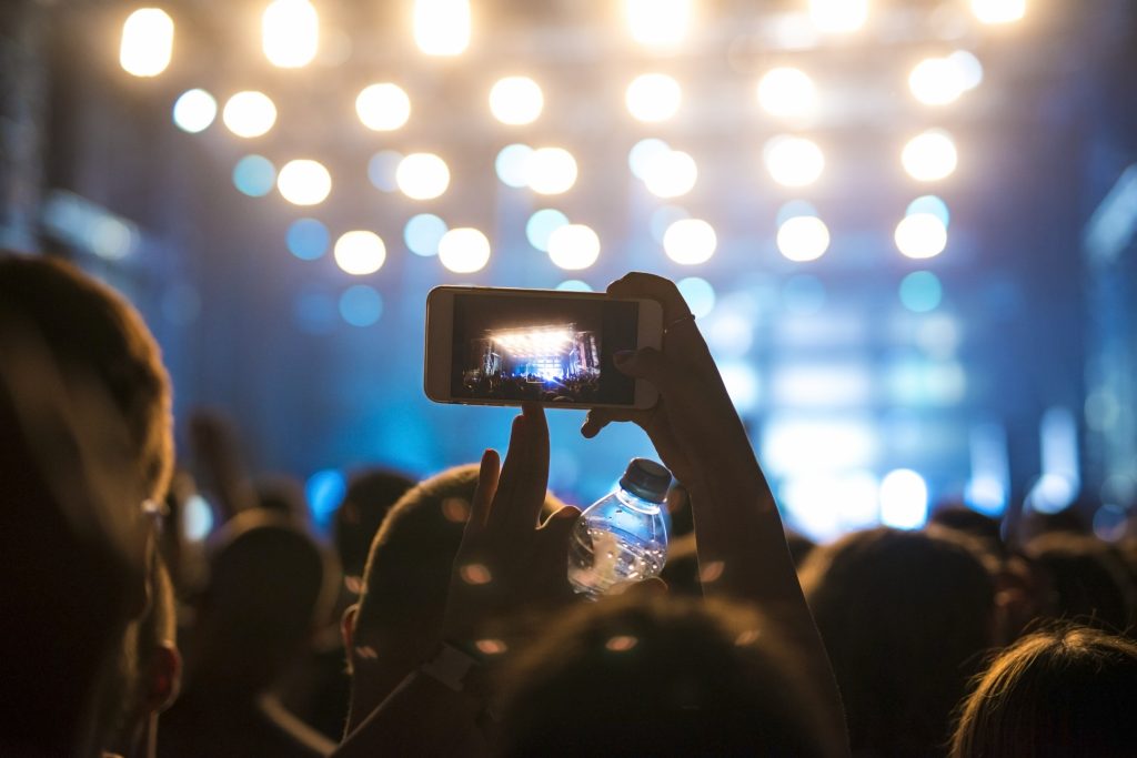Personne enregistrant un événement avec un smartphone lors d'un concert éclairé, représentant la vitalité et l'engagement dans l'industrie du divertissement.