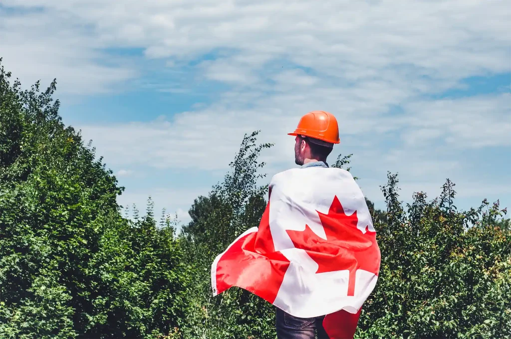 Travailleur portant un casque orange et enveloppé dans le drapeau canadien, représentant les opportunités d'emploi pour les travailleurs étrangers temporaires au Canada.